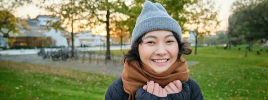 bellissimo giovane asiatico donna nel caldo cappello e sciarpa, sorrisi a telecamera, passeggiate in giro parco su freddo primavera giorno, in posa con contento viso espressione foto