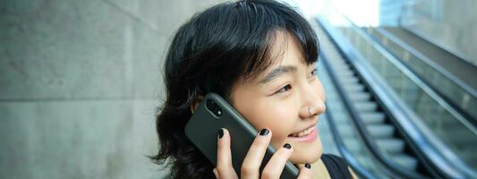 colpo alla testa di sorridente coreano donna con smartphone, fa un' Telefono chiamata, va giù scala mobile nel città, pendolari per Università foto