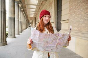 sorridente giovane testa Rossa donna nel rosso cappello, sembra a carta carta geografica per Guarda per turista attrazione. turismo e persone concetto. ragazza esplora città, provato per trova modo foto