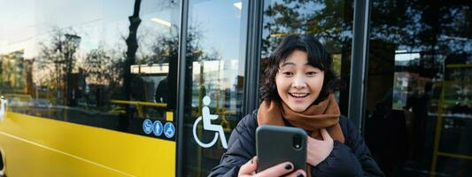 entusiasta asiatico donna, in piedi su autobus fermare con smartphone, guardare a Telefono schermo con stupito, trionfante viso, vincente, sentire grande notizia su video Chiacchierare foto