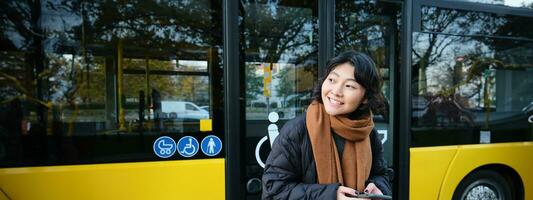 ritratto di coreano ragazza guardare per sua autobus su un' fermare, Tenere mobile Telefono, controllo orario, tempo tavolo su smartphone App foto