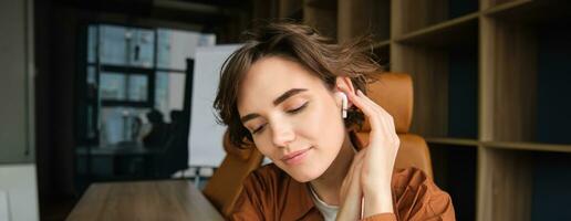 vicino su ritratto di donna nel un ufficio, ascoltando per musica nel senza fili cuffia, sorridente mentre godendo preferito canzone foto