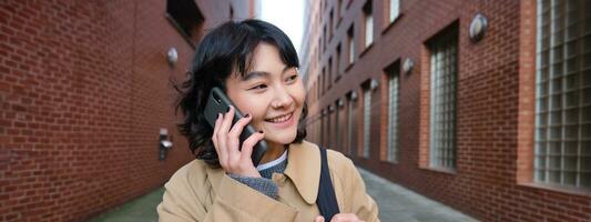 ritratto di giovane coreano donna a piedi giù strada con Telefono, parlando con qualcuno, fa un' chiamata, ha telefono conversazione foto