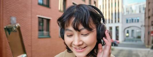 vicino su ritratto di elegante brunetta asiatico ragazza, ascolta musica nel cuffia, tocchi auricolari e sorrisi, gode preferito canzone, sta su strada foto