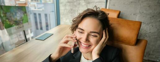 vicino su ritratto di bellissimo ufficio gestore, dipendente seduta nel sua ufficio, ridendo e sorridente durante un' Telefono chiamata, Tenere smartphone, riposo su pranzo rompere a opera foto