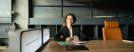 giovane donna d'affari in attesa per inizio di incontro. aziendale donna nel completo da uomo seduta nel ufficio, Lavorando su documenti, in attesa per cliente foto