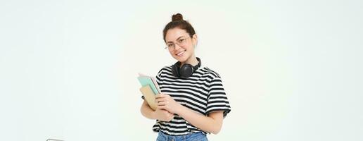 formazione scolastica concetto. sorridente brunetta ragazza, alunno nel casuale Abiti, detiene sua libri, studia Materiale, indossa cuffie al di sopra di collo, sembra fiducioso e felice, isolato al di sopra di bianca sfondo foto