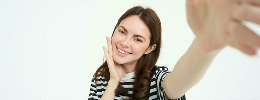 stile di vita e persone concetto. giovane spensierato donna, sorridente mentre assunzione autoscatto su smartphone, in posa per foto, in piedi al di sopra di bianca sfondo foto