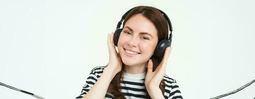 ritratto di bellissimo donna nel senza fili cuffia, ascoltando musica, utilizzando auricolari, sorridente a telecamera, in piedi al di sopra di bianca sfondo foto