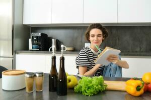 ritratto di giovane sorridente donna nel cucina, Tenere taccuino, fabbricazione Appunti per ricetta, scrittura drogheria elenco, cucinando insalata, seduta vicino verdure e chopping tavola foto