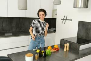 moderno contento donna danza e cucinando, ascoltando musica nel cuffia, fabbricazione un' vegano salutare pasto, vegetariano cibo foto