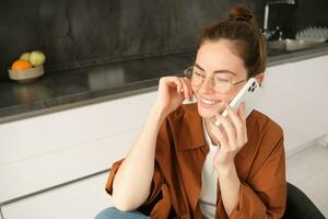 moderno giovane donna seduta nel cucina con smartphone, parlando su mobile Telefono, ha amichevole conversazione, chiamata qualcuno foto