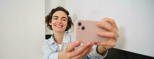 ritratto di bellissimo brunetta ragazza, prende autoscatto su smartphone nel sua cucina, in posa per foto su mobile Telefono