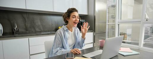 bellissimo ragazza onde mani a il computer portatile telecamera, dice Ciao per colleghi, si connette per opera incontro in linea, studi su a distanza a partire dal sua cucina foto