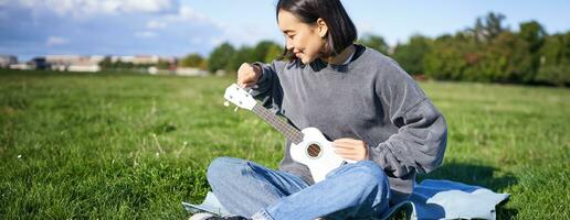 cantando asiatico ragazza giocando ukulele su erba, seduta su coperta nel parco, rilassante all'aperto su soleggiato giorno foto