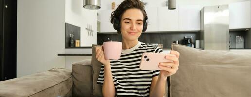 ritratto di donna nel sua vivente camera, seduta su divano e Guardando video su mobile Telefono, indossare cuffia, potabile tè o caffè nel rosa tazza foto