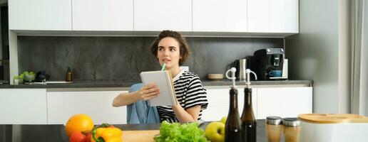 ritratto di pensiero donna con taccuino, cucinando, scrittura giù ricetta ingredienti, decidere su un' pasto per cena, seduta vicino verdure e chopping tavola nel cucina foto