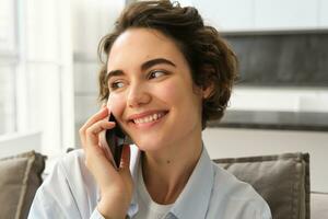 vicino su ritratto di sorridente bellissimo donna, parlando su mobile Telefono, chat su smartphone, chiamate qualcuno, si siede a casa foto