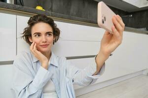 ritratto di giovane donna si siede su cucina pavimento con telefono, prende autoscatto su smartphone con App filtri, pose per foto su mobile Telefono