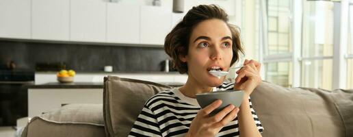 vicino su ritratto di donna mangiare nel davanti di tv, guardare stupito e con interesse a schermo, mangiare colazione, Tenere ciotola di cereali e cucchiaio, seduta su divano affascinato foto