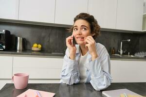 ritratto di donna pensiero mentre parlando su mobile Telefono, seduta con smartphone nel cucina a casa e pungente matita con confuso viso foto