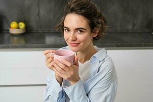 contento le mattine. ritratto di contento brunetta donna, bevande tazza di caffè nel sua cucina e sorridente, accogliente e caldo inizio di il giorno con cuppa foto