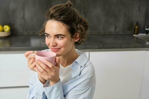 ritratto di sorridente, bellissimo giovane donna, potabile caffè nel cucina, mattina Magia con cuppa Tè, guardare teneramente foto