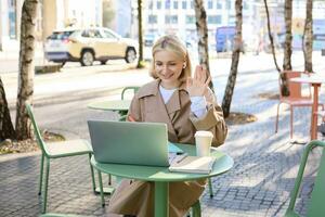 allegro ragazza con computer portatile, detto ciao, si connette per in linea incontro, agitando mano a telecamera, chat con qualcuno attraverso Internet, seduta nel all'aperto caffè negozio foto