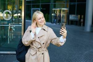 stile di vita ritratto di giovane moderno donna, carino ragazza video chat su strada, agitando mano a sua smartphone e sorridente, a piedi su strada e parlando per amico su mobile Telefono App foto