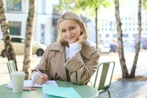 ritratto di giovane moderno donna nel elegante trincea cappotto, seduta nel all'aperto bar su soleggiato giorno, scrittura nel sua taccuino, Lavorando o fare compiti a casa, potabile caffè foto