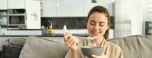 Immagine di sorridente, contento giovane donna mangiare colazione, Tenere ciotola di cereali con latte, avendo pasto a casa foto