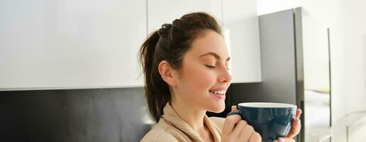 ritratto di contento giovane donna inizia sua mattina con boccale di caffè, potabile tè a partire dal tazza, in piedi nel il cucina, sorridente allegramente foto