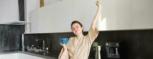 ritratto di spensierato brunetta donna, danza e allungamento mani nel il cucina, potabile caffè, sorridente lieta nel il mattina foto