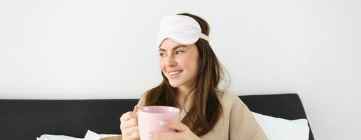 ritratto di bellissimo donna nel addormentato Borsa e pigiama, potabile caffè nel letto, godendo sua luminosa, rilassante mattina foto