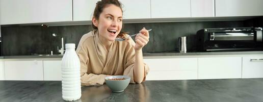 ritratto di giovane bellissimo donna nel accappatoio, mangiare cereali per colazione, Leans su cucina piano di lavoro, guardare a sua mattina pasto foto