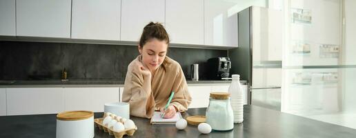 attraente giovane allegro ragazza cottura al forno a il cucina, fabbricazione Impasto, Tenere ricetta prenotare, avendo idee foto