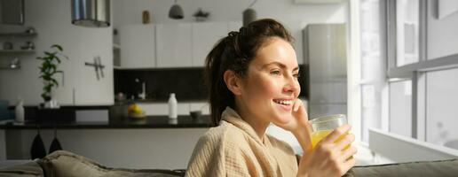 vicino su di bellissimo giovane donna su divano, bevande arancia succo e sorrisi, sembra al di fuori finestra, gode mattina a casa foto