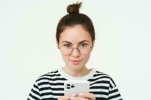 ritratto di bellissimo giovane donna nel occhiali, Tenere smartphone, utilizzando mobile Telefono, in linea shopping nel applicazione, in piedi al di sopra di bianca sfondo foto