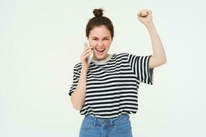 entusiasta ragazza cantando, risposte Telefono chiamata e sembra emozionato, trionfante al di sopra di grande notizia, sta al di sopra di bianca sfondo foto