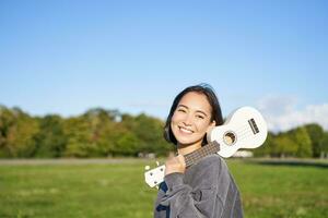 giovane fricchettone ragazza, viaggiatore Tenere sua ukulele, giocando all'aperto nel parco e sorridente foto