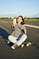 positivo adolescente asiatico ragazza prende autoscatto, video chat su mobile Telefono app, si siede su skateboard e record video blog foto