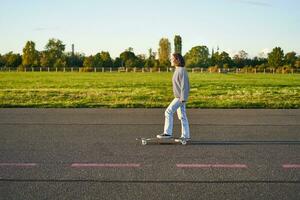 bellissimo asiatico pattinatore ragazza equitazione sua longboard su soleggiato vuoto strada. giovane donna godendo sua pattinare cavalcata sorridente e ridendo foto