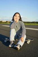 verticale tiro di asiatico ragazza pattinatore, si siede su sua skateboard e sorrisi, gode soleggiato giorno, crociera su longboard su vuoto strada all'aperto foto