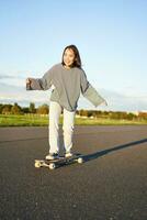 carino asiatico ragazza equitazione skateboard, pattinando su strada e sorridente. pattinatore su incrociatore longboard godendo all'aperto su soleggiato giorno foto