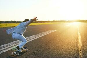 contento pattinatore ragazza equitazione sua skateboard e avendo divertimento su vuoto strada. sorridente donna godendo incrociatore cavalcata su soleggiato strada foto