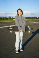 verticale tiro di pattinatore ragazza in posa con longboard, crociera su vuoto strada nel periferia. sorridente asiatico donna pattinando su skateboard, Tenere incrociatore nel mani foto
