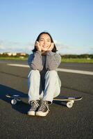 verticale tiro di asiatico ragazza pattinatore, si siede su sua skateboard e sorrisi, gode soleggiato giorno, crociera su longboard su vuoto strada all'aperto foto