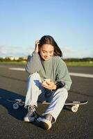 verticale tiro di asiatico donna seduta su skateboard su strada, Tenere smartphone app. pattinatore ragazza pattini su longboard, utilizzando mobile Telefono foto