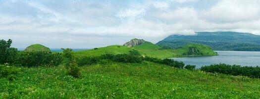 naturale paesaggio di kunashir isola con erboso colline, vulcanico rocce, vulcano nel il nuvole e un' valle con un' laguna foto