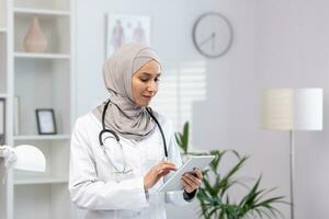 musulmano donna nel bianca medico cappotto in piedi dentro medico ufficio di clinica, femmina medico utilizzando tavoletta computer pensiero e concentrando, concentrato donna a opera nel Ospedale foto